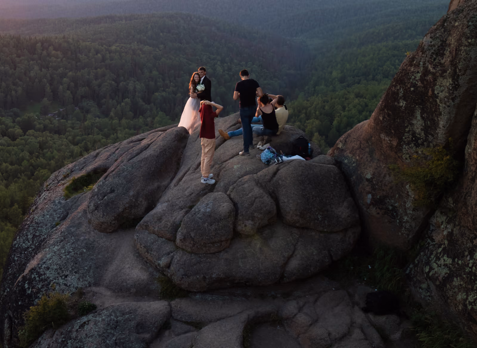 A photographer on a rock outcrop at dusk photographing a bride and groom above a forested valley